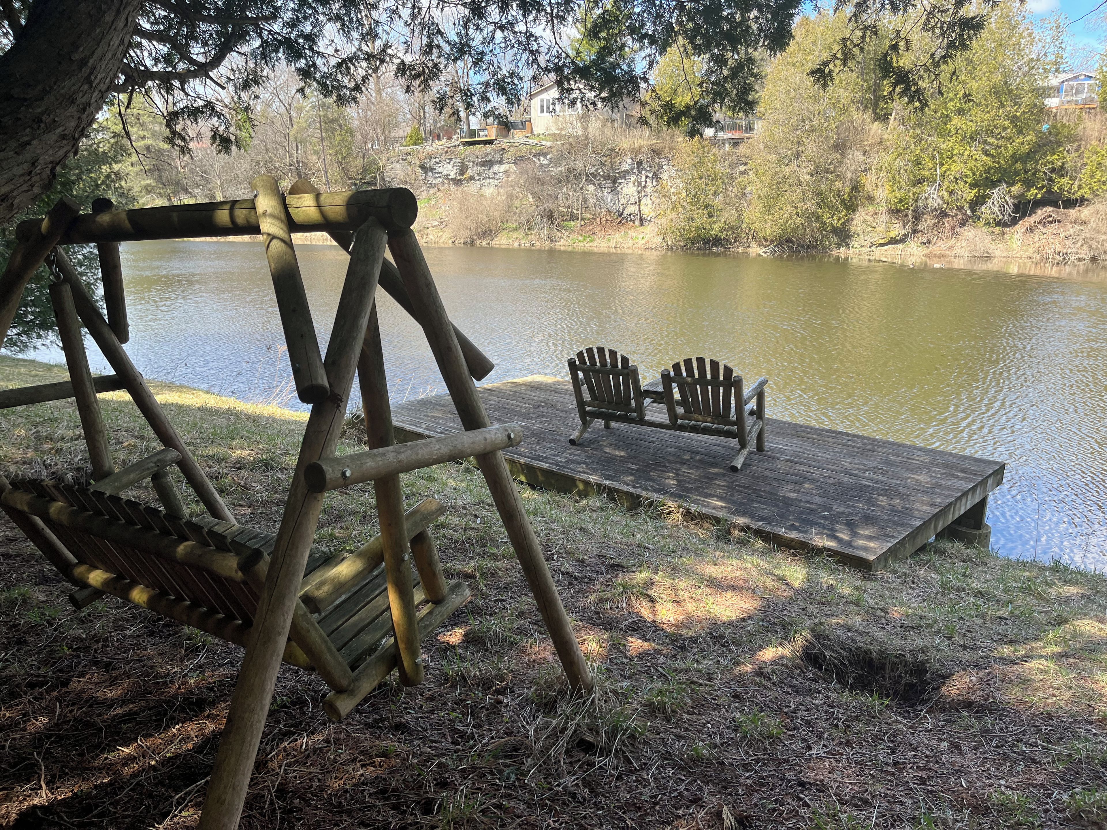 A wooden swing set and Adirondack chairs by a calm lake with a dock.