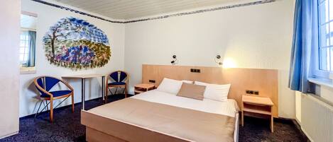 A hotel room with a large bed, two bedside tables, a desk with two chairs, a wall painting, and a window with curtains.