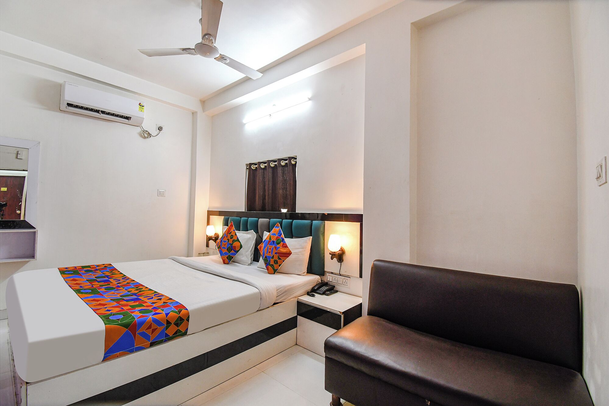 A hotel room with a bed, a bedside table with a phone, a chair, a ceiling fan, an air conditioning unit, and a wall-mounted light fixture.