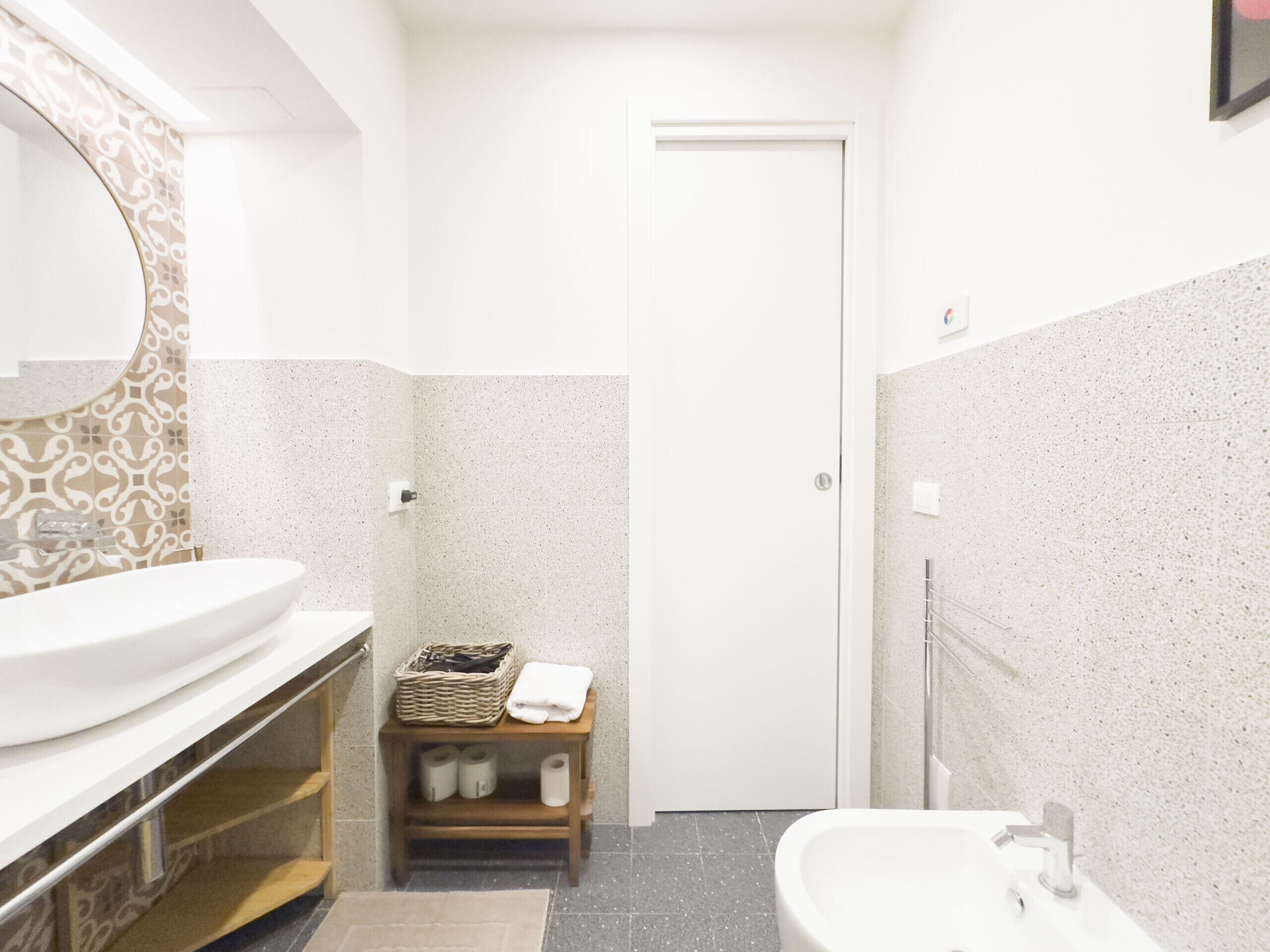 Une salle de bain avec un lavabo blanc, un miroir, une petite étagère en bois où sont posées des serviettes et des articles de toilette, et une porte qui donne sur une autre pièce.