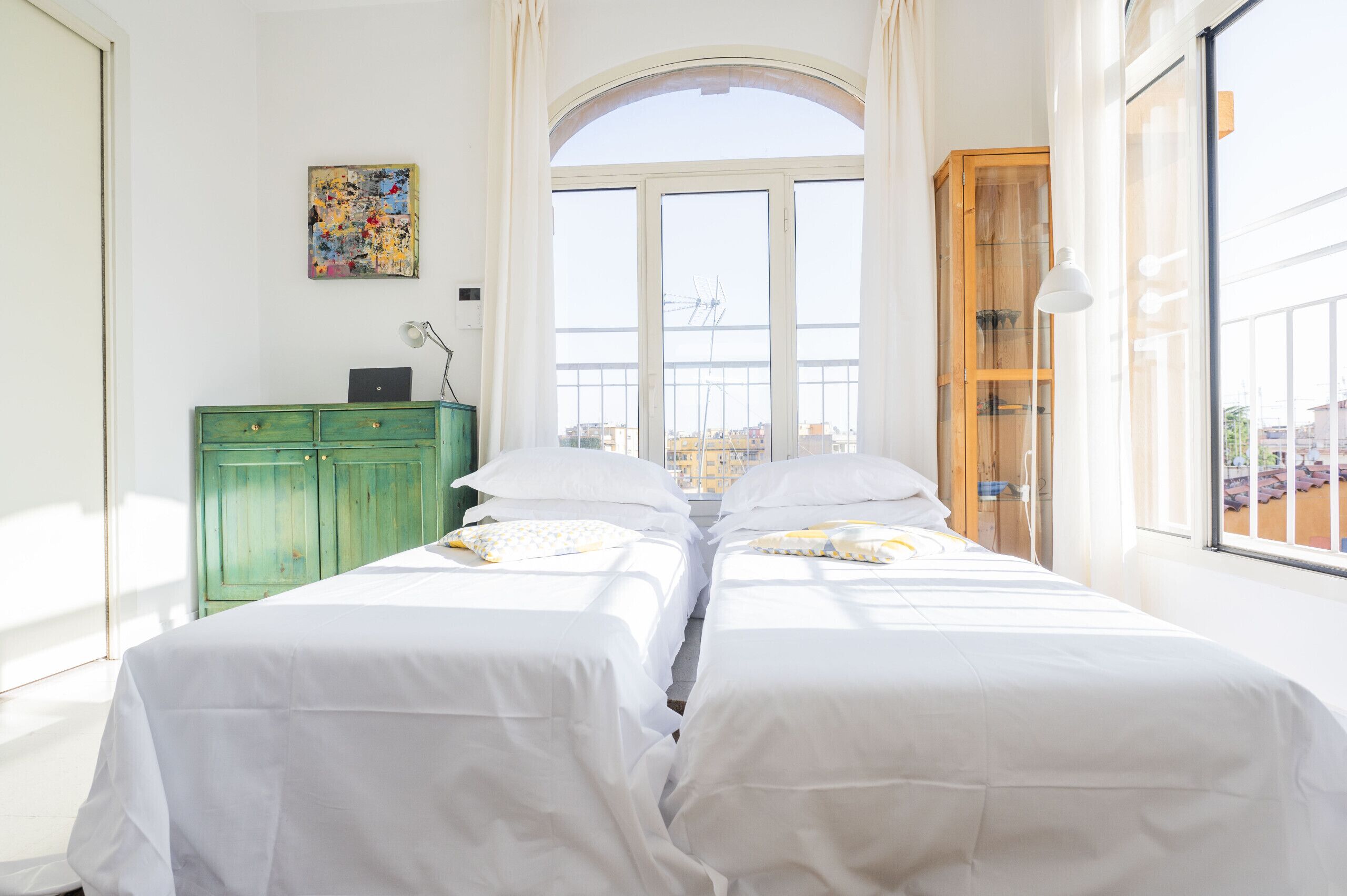 Une chambre avec deux lits, une commode verte, un tableau au mur et une fenêtre donnant sur des immeubles.