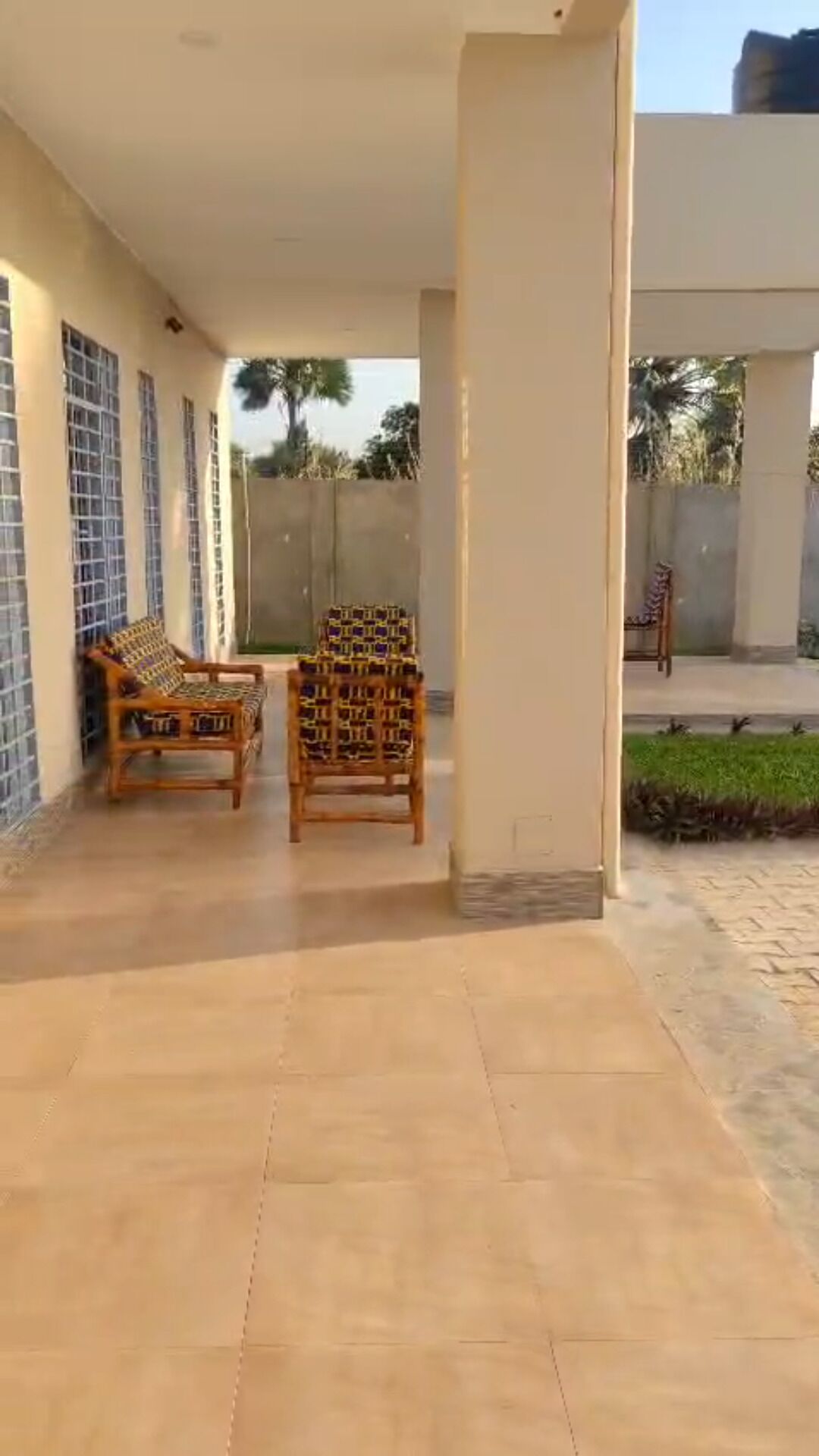 A covered patio area with two wooden chairs and a sofa, a tiled floor, and a view of a garden and building in the background.