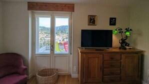 A room with a television on a wooden cabinet, a wicker basket, a red chair, and a view of a town through a door.
