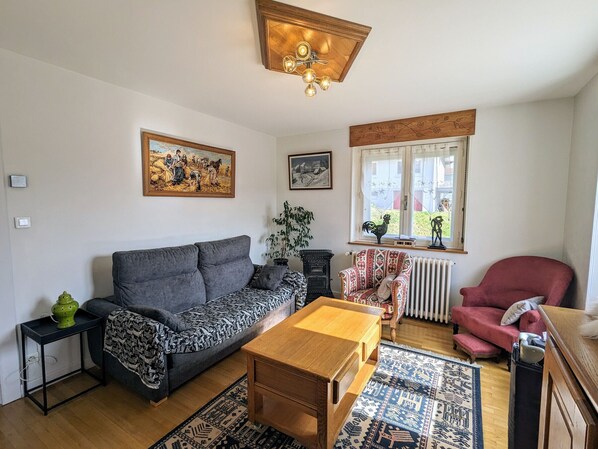 A living room with a grey sofa, a patterned armchair, a wooden coffee table, and a red armchair. There are two side tables, one with a green lamp and the other with a black lamp. A large painting and a smaller framed picture adorn the walls.