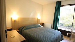 A hotel room with a bed, bedside tables, a nightstand, a window with curtains, and a radiator.