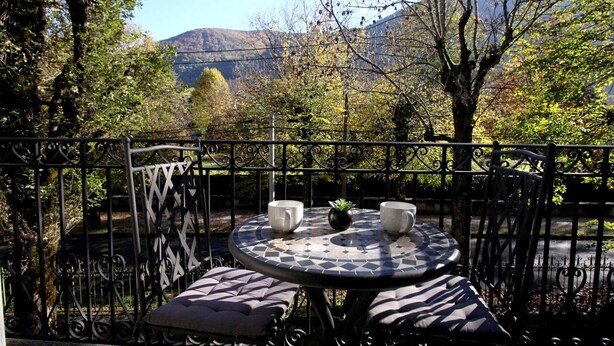A round table with two white cups and a potted plant, surrounded by a black wrought iron fence with a view of trees and mountains in the background.