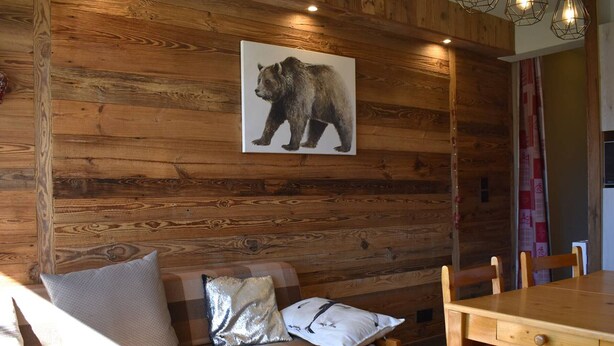 Een houten wand met een beren afbeelding, een houten bank met kussens, en een houten eettafel met stoelen.