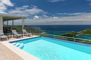 A pool with a view of the ocean, lounge chairs, and a covered area.