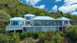 A large, multi-story beachfront property with a pool, multiple balconies, and lush greenery.