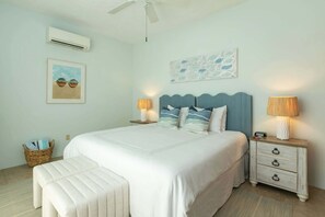 A bedroom with a large bed, two bedside tables with lamps, a wall-mounted air conditioner, a framed picture, and a decorative wall piece.