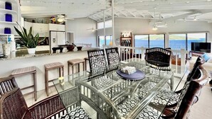 A spacious living area with a glass-top dining table surrounded by patterned chairs, a high ceiling with exposed beams, and a view of the ocean through large windows. - Oceanfront Villa-Private Pool-Panoramic Views -Sleeps 12 (St. Thomas)