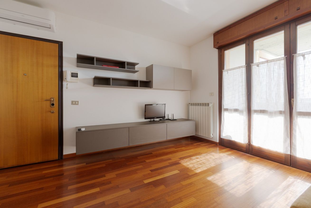 A room with a wooden floor, a television on a grey cabinet, a door, and a window with curtains.