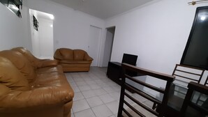 A living room with a brown leather sofa, a wooden dining table with chairs, and a black TV stand. - Round trip - LA26-Bertioga, SESC, Riviera, Vista Linda, Indaiá (Jardim Rafael Bertioga)