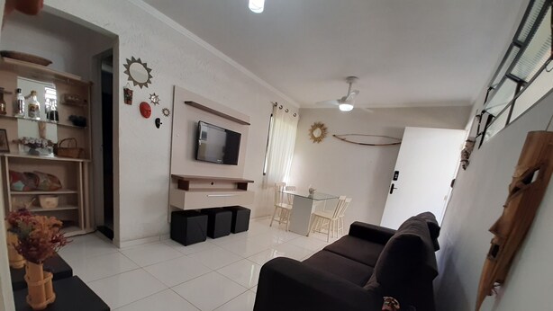 A living room with a black sofa, a TV mounted on the wall, a dining table with chairs, and a small kitchen area. - Round trip - LA26-Bertioga, SESC, Riviera, Vista Linda, Indaiá (Jardim Rafael Bertioga)
