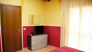 A bedroom with a wooden door, a dresser with a TV, a bed with a red bedspread, and a window with curtains.