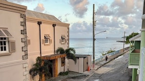 A street with a car parked on the side, a building with a balcony, and a view of the ocean.
