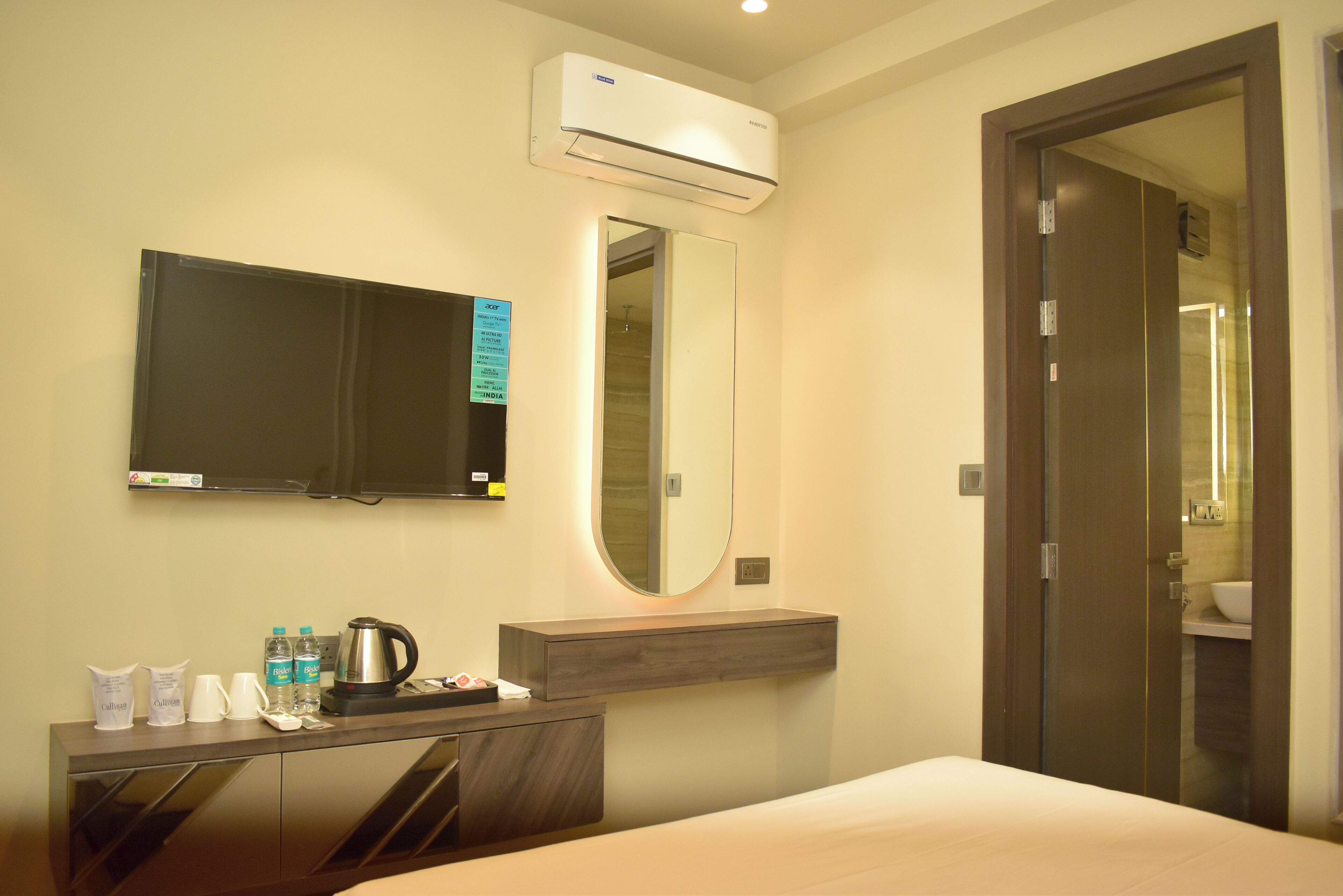 A hotel room with a bed, a bedside table with toiletries, a mirror, an air conditioning unit, and a television.