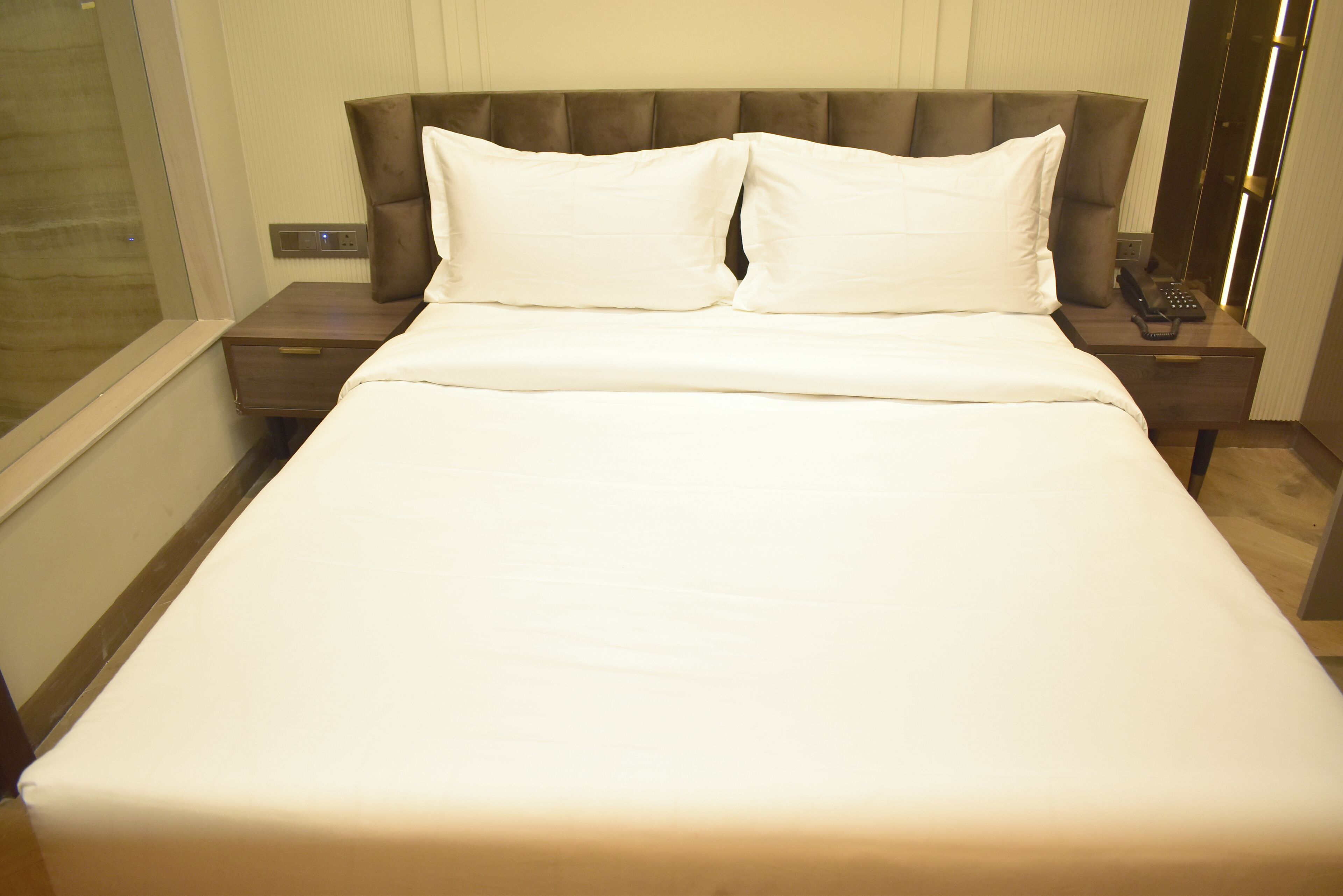 Een keurig opgemaakt bed met witte lakens, een hoofdbord met een gestoffeerde vormgeving, en een nachtkastje met een telefoon en een afstandsbediening.