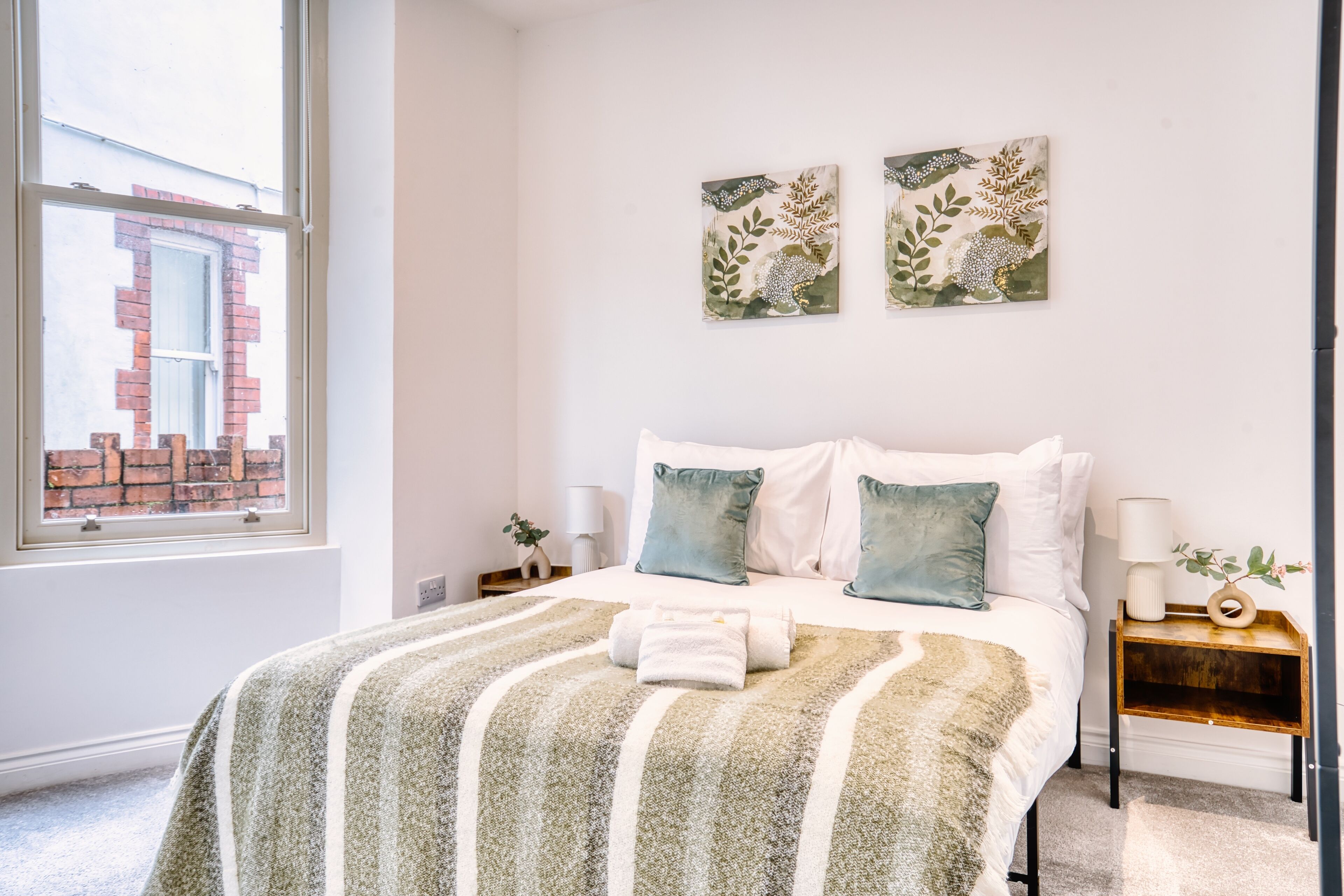 Quarto com uma janela ampla, uma cama com manta listrada, duas almofadas, uma mesa de cabeceira com abajur e duas obras de arte florais penduradas na parede.