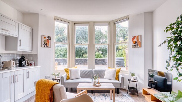 Ein modernes Wohnzimmer mit Sofa, Couchtisch und Fernseher. Es gibt eine Küchenzeile mit Schränken und ein Fenster mit Blick auf Grünflächen.