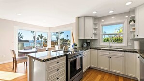 A modern kitchen with white cabinets, a granite countertop, and a large window overlooking a balcony with a view of palm trees. - Oceanview Townhome Steps from Iconic Windansea Beach (San Diego)