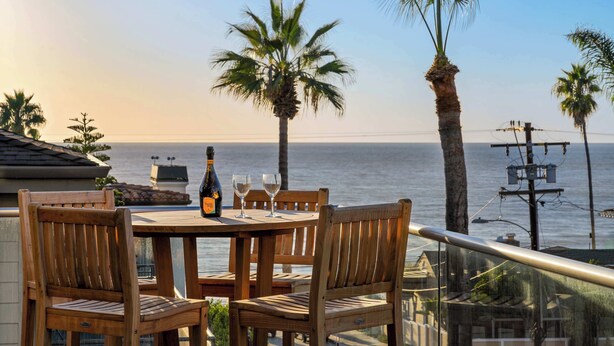 A wooden table with two chairs and a bottle of champagne, overlooking the sea with palm trees. - Oceanview Townhome Steps from Iconic Windansea Beach (San Diego)
