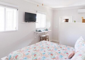 A bedroom with a bed, a TV mounted on the wall, a small table with stools, and a shelf with kitchen items. - The Palms Hideaway (Nassau)