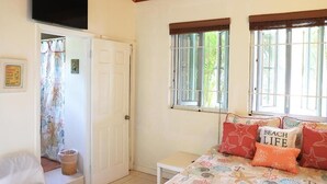 Une chambre avec un lit, un téléviseur fixé au mur, une fenêtre donnant sur un espace verdoyant et une porte menant à une autre pièce.