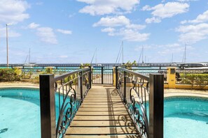 A wooden deck leading to a pool with a metal railing, surrounded by a clear sky and a distant view of boats. - Elegancia Ocean View with Cooling Pool (Kralendijk)