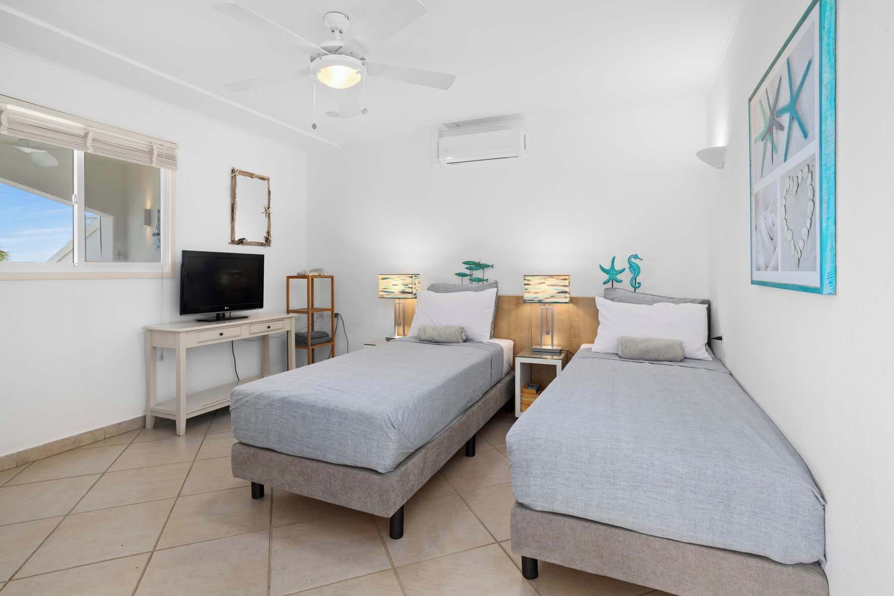 A bedroom with two single beds, a desk with a TV, a ceiling fan, and artwork on the walls.