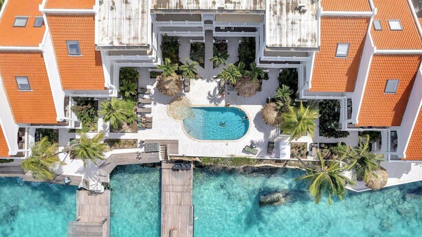 Vue aérienne d’un complexe hôtelier doté d’une piscine centrale, d’une terrasse en bois et d’une eau turquoise et cristalline.