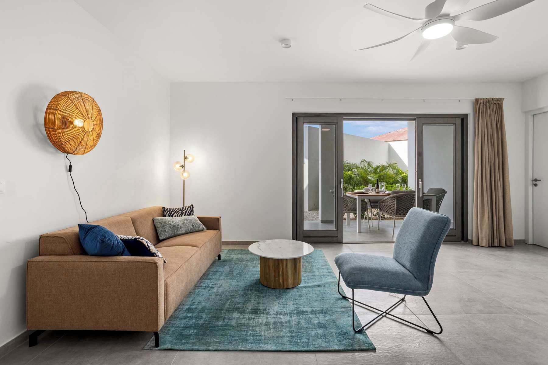 A modern living room with a sofa, a coffee table, and a chair. There is a ceiling fan, a wall-mounted light, and a sliding glass door leading to an outdoor area with a table and chairs.