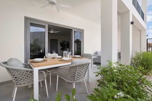 Un espace de restauration en plein air avec une table en bois et des chaises en osier, un ventilateur de plafond et une vue sur le jardin.