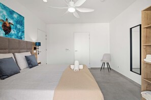 A modern bedroom with a large bed, a ceiling fan, a bedside table with a lamp, a chair, and a wooden shelf with towels.