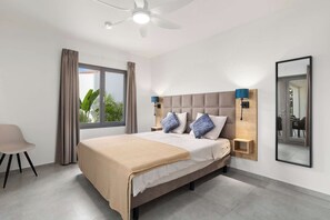 Une chambre moderne avec un grand lit, un ventilateur de plafond, une fenêtre avec des rideaux, une table de chevet avec des lampes et un miroir au mur.