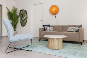 A modern living room with a light blue chair, a round wooden coffee table, a beige sofa with patterned cushions, a large mirror, and a wall-mounted orange light fixture.