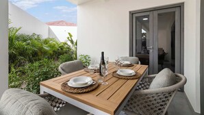 A dining area with a wooden table set for four, surrounded by wicker chairs, and a view of a red-roofed building through a glass door. - Bonaire Beach Apartments 2 with a palm garden, a refreshing shared pool and a private sitting area (Kralendijk)