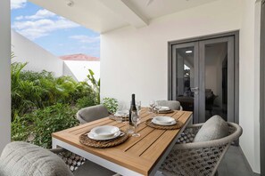 Un coin repas avec une table en bois dressée pour quatre personnes, entourée de chaises en osier, et une vue sur un bâtiment au toit rouge, visible à travers une porte vitrée.