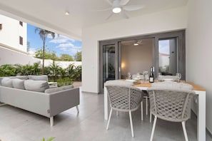 A modern outdoor dining area with a table and chairs, a sofa, and a view of a building and greenery.