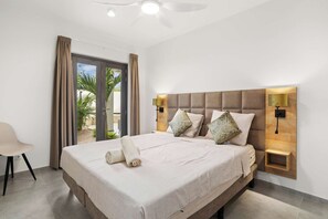 A hotel room with a large bed, two wall-mounted lamps, a ceiling fan, and a view of palm trees through the window.