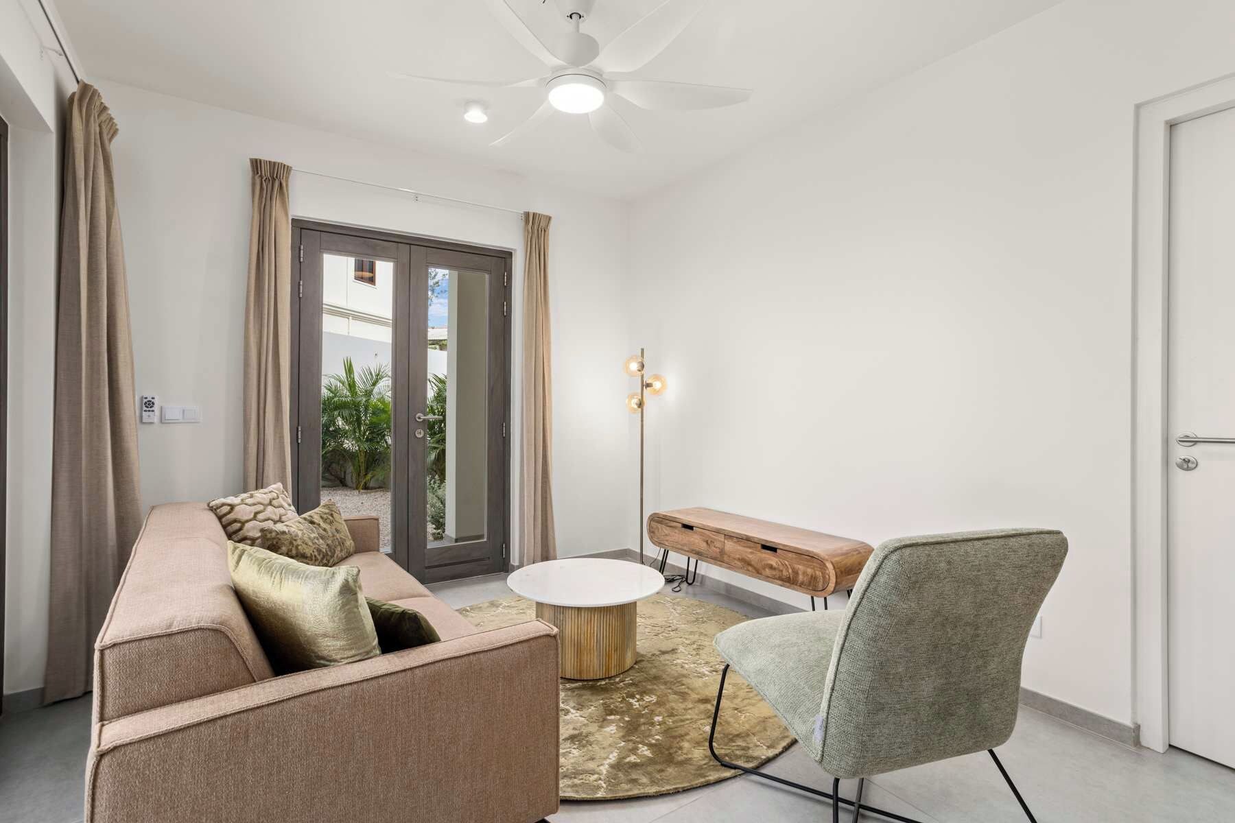 Un salon moderne comprenant un canapé, une table basse ronde, un banc et un fauteuil. Une porte donne accès à un espace extérieur agrémenté de plantes.
