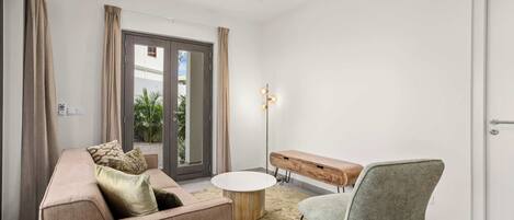 Un salon moderne comprenant un canapé, une table basse ronde, un banc et un fauteuil. Une porte donne accès à un espace extérieur agrémenté de plantes.