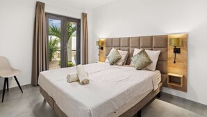 A hotel room with a large bed, two wall-mounted lamps, a ceiling fan, and a view of palm trees through the window.