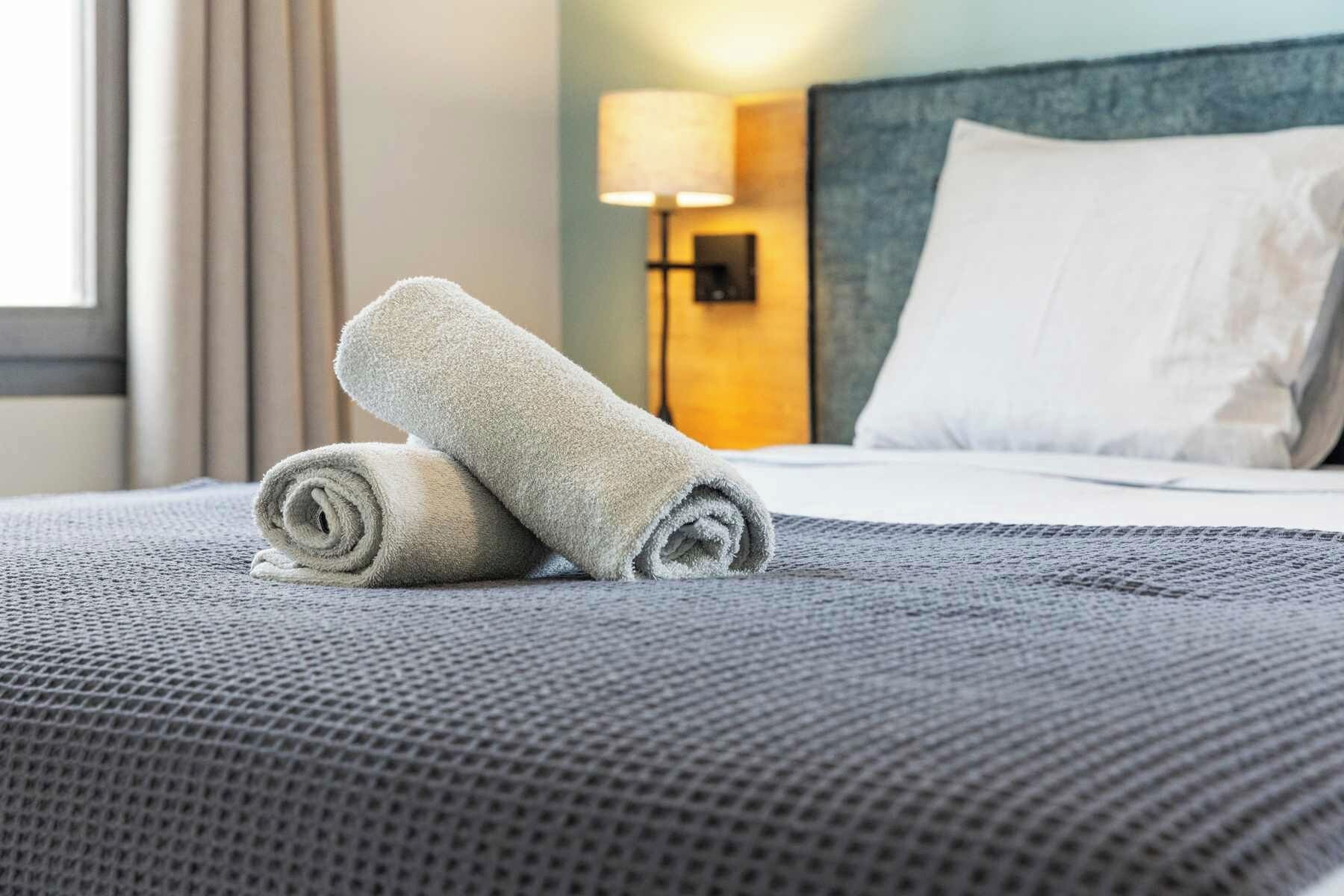 Una cama bien hecha con una colcha gris texturizada y dos toallas enrolladas.