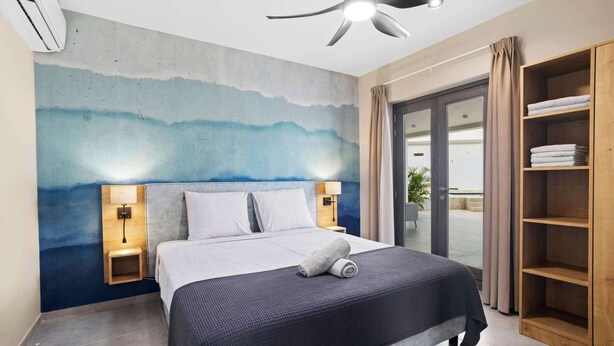 Una habitación de hotel moderna con una cama, ventilador de techo, un mural en la pared, una mesita de noche con lámpara y un estante de madera empotrado con toallas.