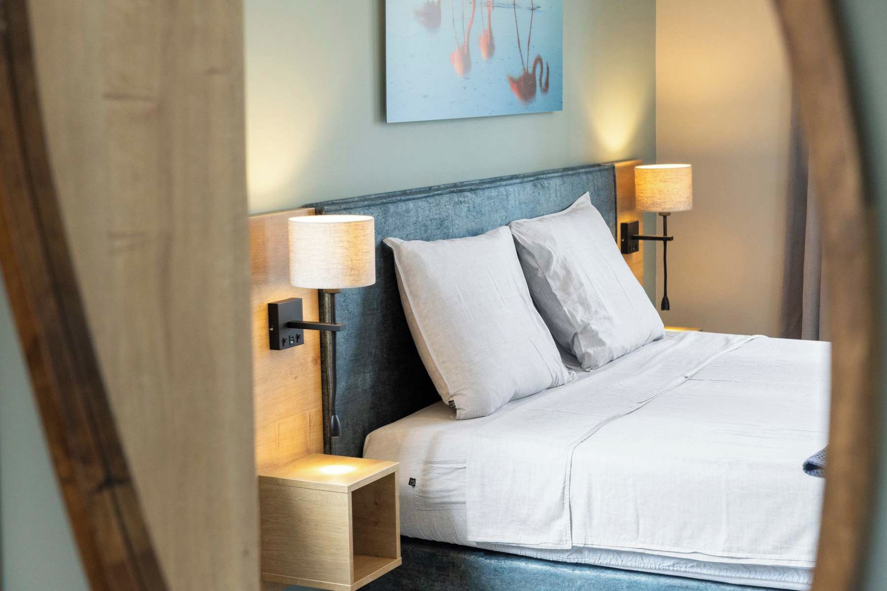 A neatly made bed with white linens and two pillows, flanked by two bedside tables with lamps, in a room with a blue accent wall and a painting above the bed.