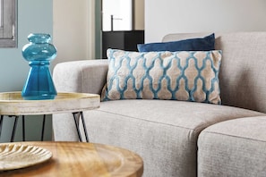 Ein Wohnzimmer mit einem beigefarbenen Sofa, einem gemusterten Kissen, einer blauen Vase und einem kleinen runden Tisch mit Holzfuß.