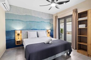 Een moderne hotelkamer met een bed, een plafondventilator, een wanddecoratie, een nachtkastje met lamp en een ingebouwde houten plank met handdoeken.