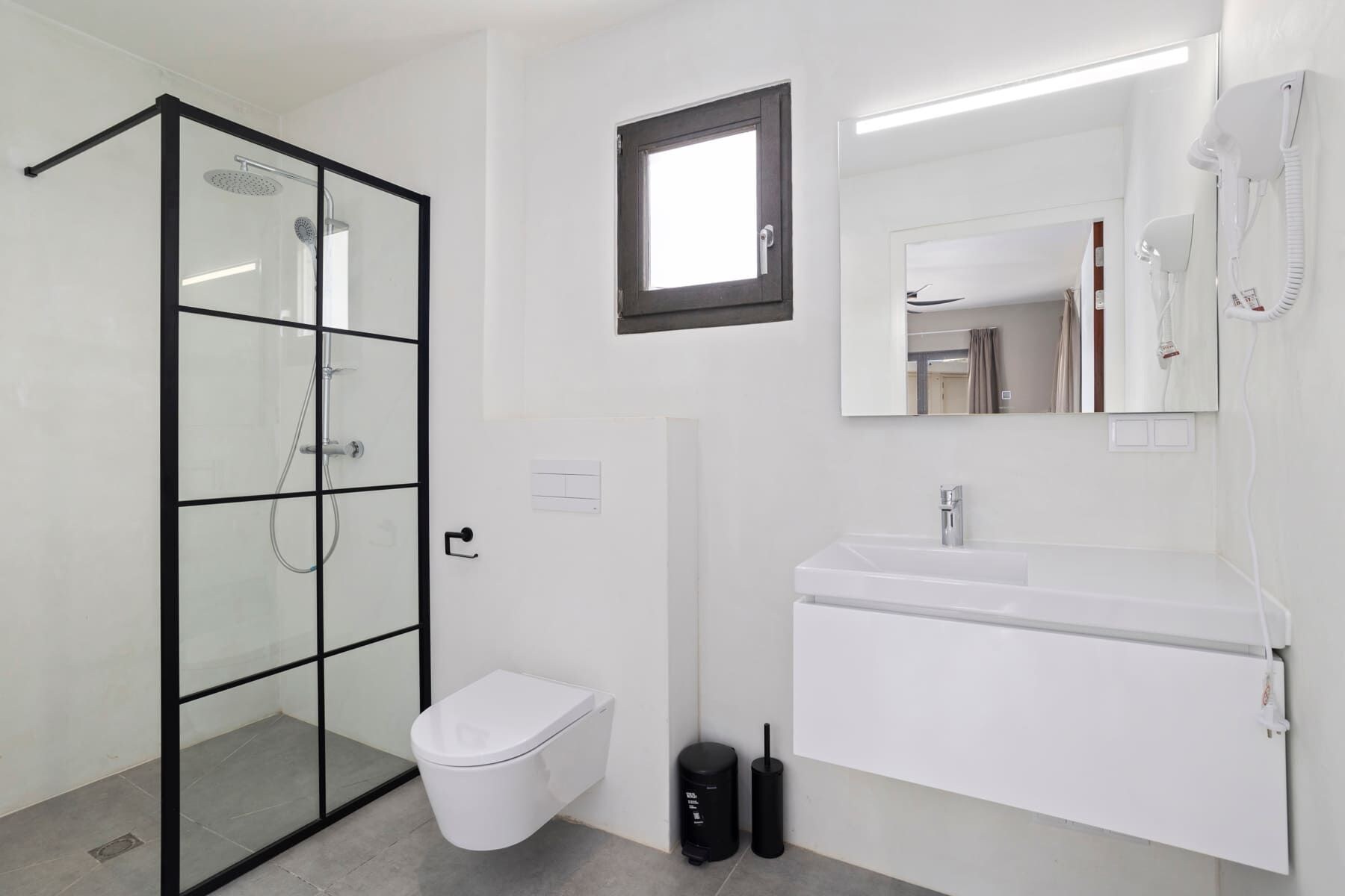 Un baño moderno con ducha de vidrio, inodoro blanco, lavamanos con espejo y secador de pelo en la pared.