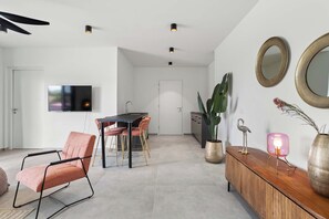 Un salon moderne comprenant un coin repas, un téléviseur à écran plat fixé au mur, un meuble en bois agrémenté d’objets décoratifs et une grande plante en pot.